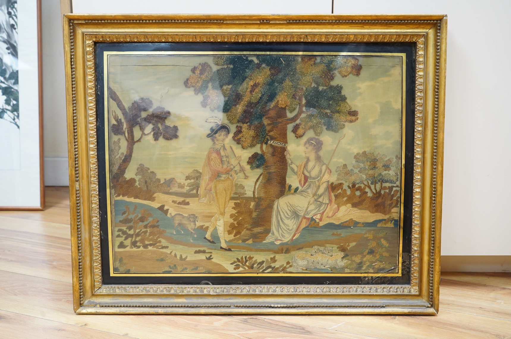 A gilt framed early 19th century silk chenille embroidered and hand painted figurative pastoral scene, framed with black and gold eglomise glass. The embroidery worked in a variety of stitches including stem and knotting